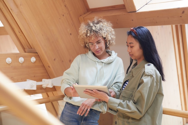 Foto due donne in piedi sulla scala in legno e utilizzando la tavoletta digitale stanno facendo un lavoro online