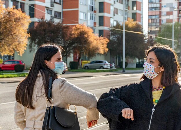 写真 コロナウイルス流行中にマスクをかぶって街を歩く2人の女性が肘を叩いて握手している