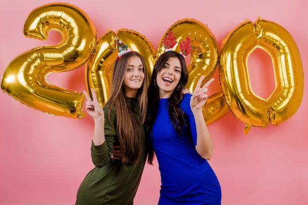 Two women wearing christmas hoop and hugging in front of golden 2020 balloons isolated over pink