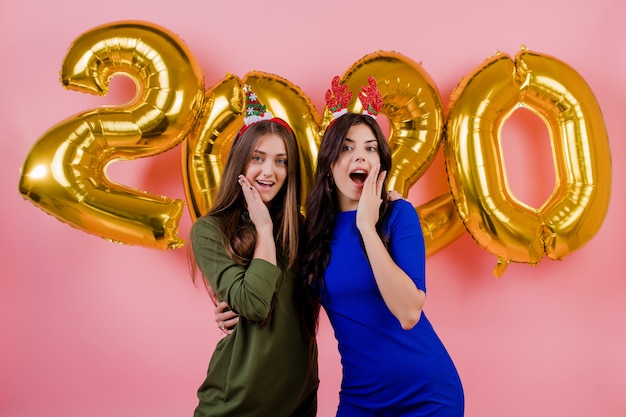 Two women wearing christmas hoop and hugging in front of golden 2020 balloons isolated over pink