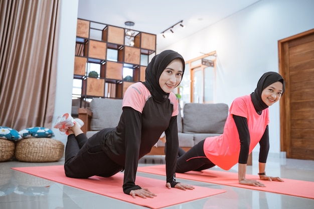 Two women wearing hijab sportswear smile while doing push ups together on a mat on the floor in the house