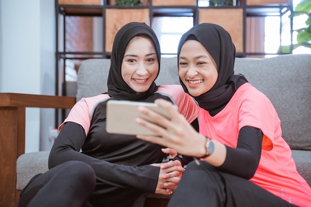 Two women wearing hijab sportswear smiling while taking selfies together with a cellphone while sitting on the floor in the house