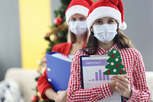 Two women wearing medical protective masks and santa claus hats holding folders with documents and small christmas tree. Business annual report on coronavirus pandemic concept
