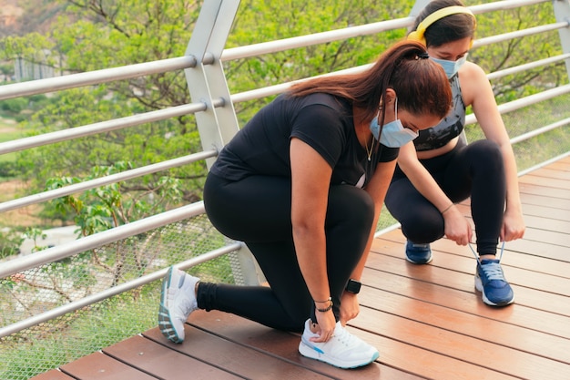 Two women wearing protective masks tie their shoelaces in preparation for a run.