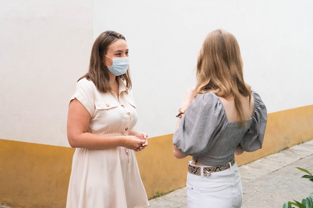 Two women wearing a surgical mask and talking on a city street during pandemic