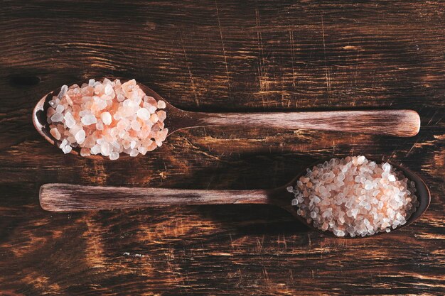 Two wooden spoons lying on a wooden background are filled with pink salt