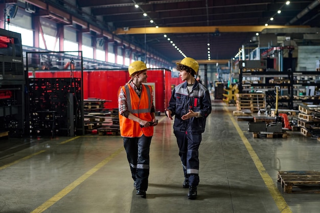 Two workers of modern factory walking down long and wide aisle and talking