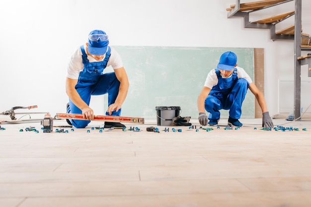 Two workers in a special uniform laying tiles with tile leveling system and laser level on the floor