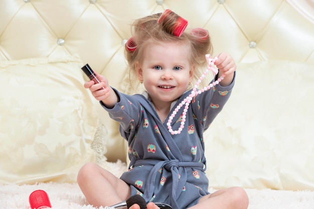 Foto bambina di due anni. una bambina in bigodini si siede sul letto in camera da letto con cosmetici e gioielli. preparazione della festa.