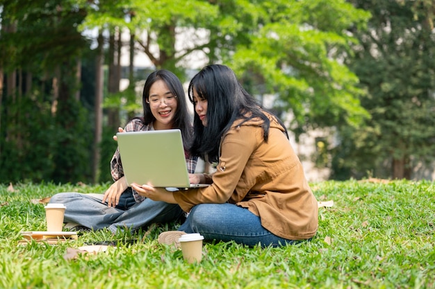 Two young Asian female students are sitting on the grass in a park studying together with a laptop