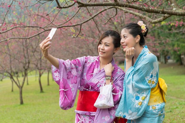 Two young asian females taking selfie using their cell phone under the cherry blossom tree. one holding cell phone another put her hands under the cheek