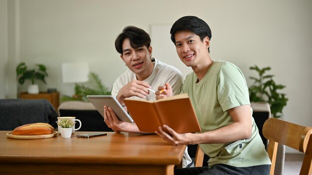 写真 カメラを見て、デジタル タブレットと本を持ったテーブルに座っている 2 人の若いアジア人男性