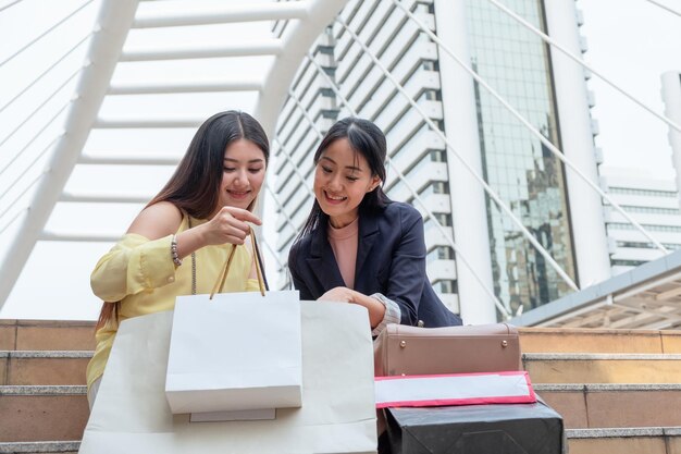 Two young asian woman sitting and showing shopping bag of purchases with friend on stair in the city