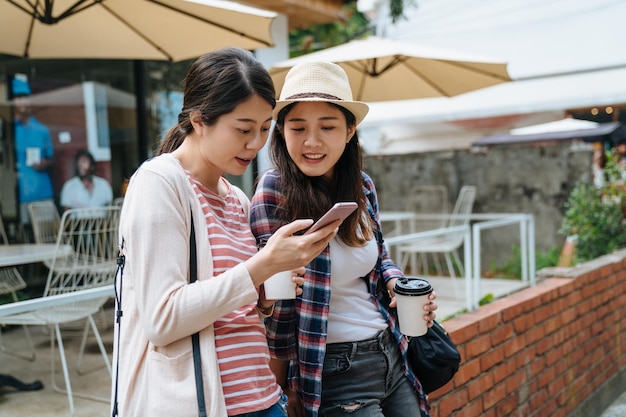 Two young asian women sit lean on red brick wall in outdoor\
cafe bar garden drink coffee in cups enjoying sunny day spring.\
girl friends relax on sunshine using smartphone searching online\
map app.