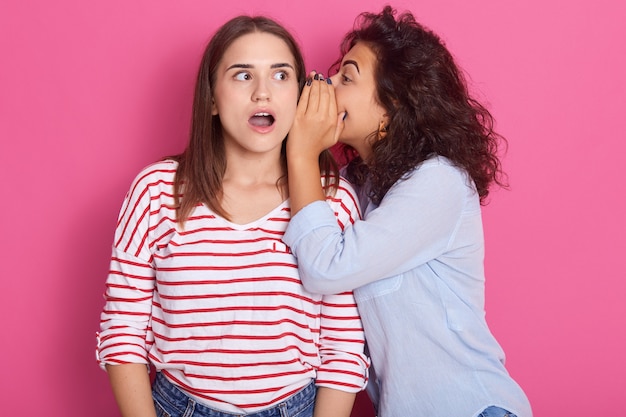 Photo two young attractive european girls wearing casual clothes standing and posing isolated over pink wall, studio portrait. people lifestyle concept. woman wispering secret to friend's ear.