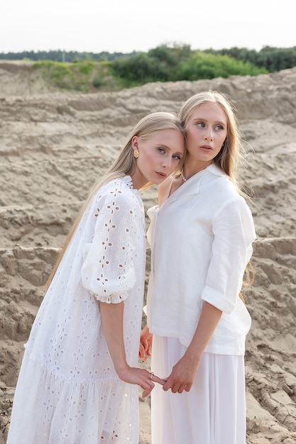 Two young attractive twins with long hair spending time together outdoors  at summer, posing at sand quarry in white dress, skirt, jacket