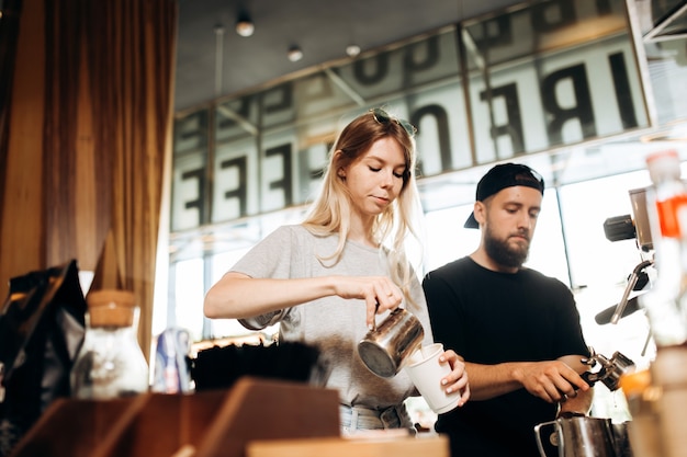 Due giovani baristi, una ragazza bionda e un uomo elegante con la barba, vengono mostrati mentre cucinano il caffè insieme in una macchina da caffè in un'accogliente caffetteria. .