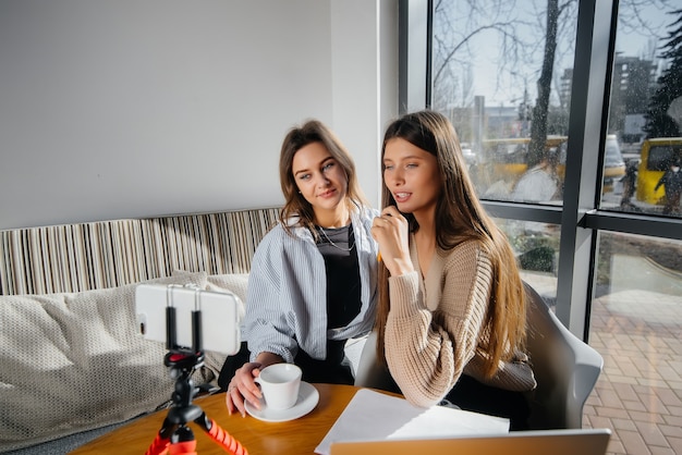 두 명의 아름다운 소녀가 카페에 앉아 비디오 블로그를 녹화하고 소셜 네트워크에서 소통하고 있습니다.