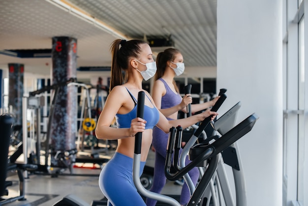 Two young beautiful girls exercise in the gym wearing masks during the pandemic