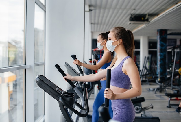 Two young beautiful girls exercise in the gym wearing masks during the pandemic