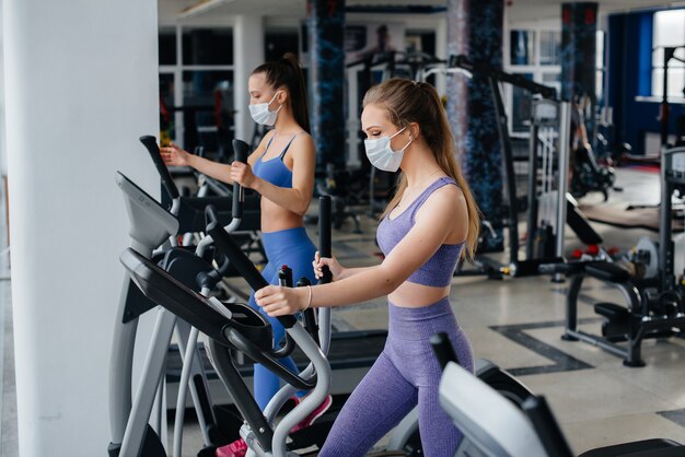 Two young beautiful girls exercise in the gym wearing masks during the pandemic