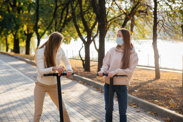 暖かい秋の日に、マスクをした2人の若い美しい女の子が公園で電動スクーターに乗ります。公園で散歩する。