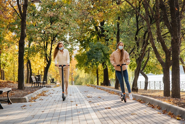 Due giovani belle ragazze in maschera cavalcano scooter elettrici nel parco in una calda giornata autunnale. camminare nel parco.