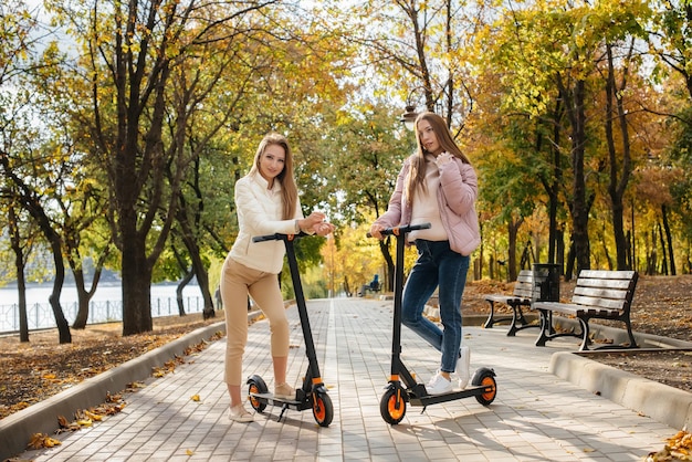 두 명의 아름다운 소녀가 따뜻한 가을날 공원에서 전기 스쿠터를 타고 있습니다.