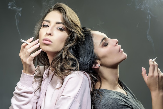 회색 배경에서 담배를 피우는 두 명의 아름다운 소녀들이 젊은 사람들의 유해하고 나쁜 습관