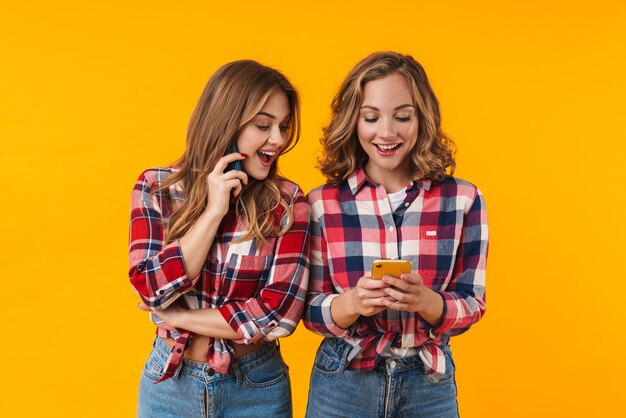 two young beautiful girls wearing plaid shirts smiling and using cellphones isolated