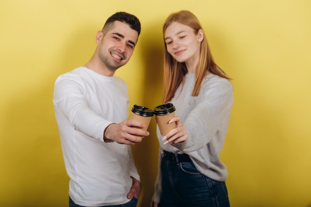 Two young beautiful people girl and guy standing on a yellow background They drink coffee