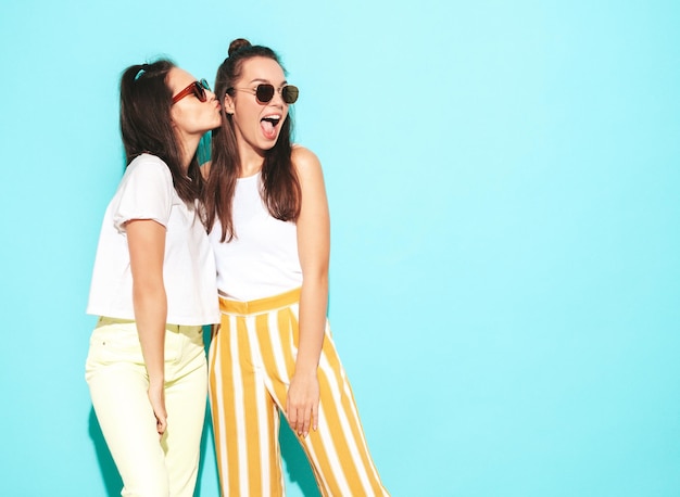 Two young beautiful smiling blond hipster female in trendy summer clothes Sexy carefree women posing near blue  wall in studio Positive models having fun Cheerful and happy in sunglasses