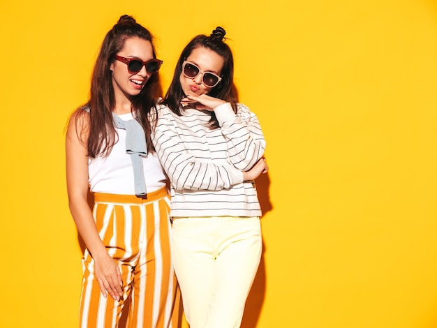 Two young beautiful smiling blond hipster female in trendy summer clothes Sexy carefree women posing near yellow wall in studio Positive models having fun Cheerful and happy in sunglasses