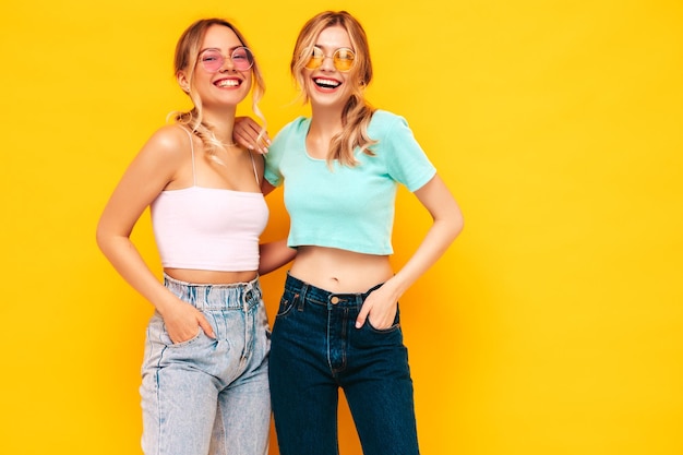 Two young beautiful smiling blond hipster female in trendy summer clothes Sexy carefree women posing near yellow wall in studio Positive models having fun Cheerful and happy In sunglasses