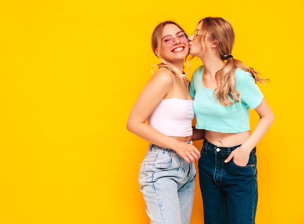 Two young beautiful smiling brunette hipster female in trendy summer clothes Sexy carefree women posing near yellow wall in studio Positive models having fun Cheerful and happy Kissing her friend