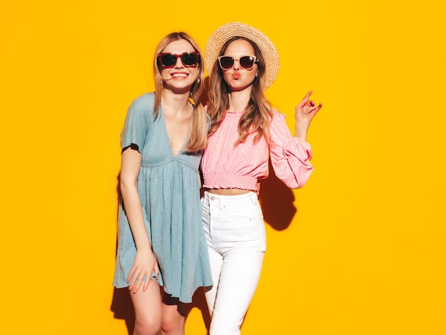 트렌디 한 여름 드레스에 두 젊은 아름 다운 미소 갈색 머리 힙 스터 여성 노란색 벽 근처 포즈 섹시 한 평온한 여성 긍정적인 모델 재미 명랑 하 고 행복 한 모자와 선글라스
