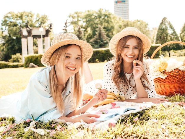 Foto due giovani belle donne sorridenti in prendisole e cappelli estivi alla moda. donne spensierate che fanno picnic all'esterno.