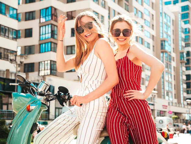 Two young beautiful smiling hipster female in trendy overallssexy carefree women driving retro motorbike on the street backgroundpositive models having fun riding classic italian scooter in eyewear