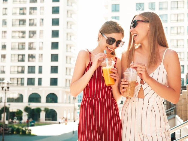 Two young beautiful smiling hipster female in trendy summer clothes Carefree women posing outdoorsPositive models holding and drinking fresh cocktail smoothie drink in plastic cup with straw