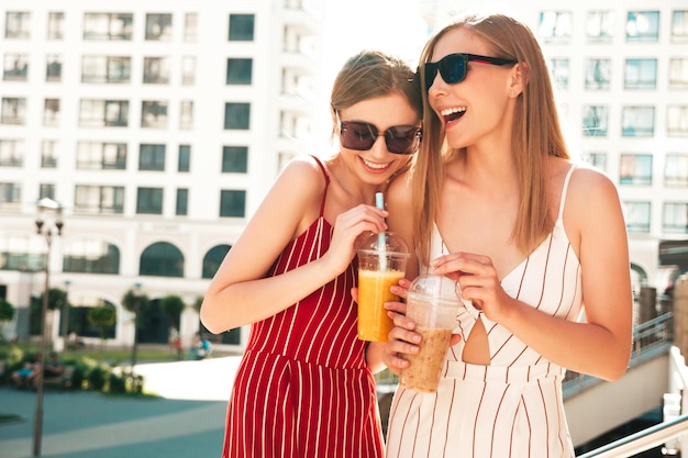 Two young beautiful smiling hipster female in trendy summer clothes Carefree women posing outdoorsPositive models holding and drinking fresh cocktail smoothie drink in plastic cup with straw