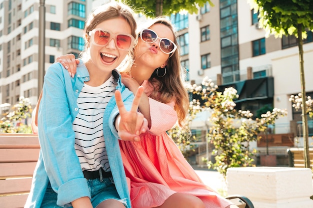 Two young beautiful smiling hipster female in trendy summer clothes and dressSexy carefree women posing in the street Positive pure models having fun at sunset sitting on a bench