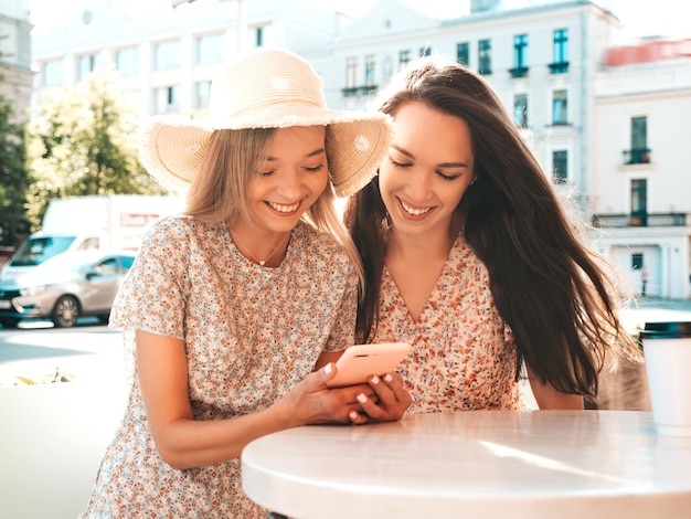 Due giovani e belle donne hipster sorridenti in abiti estivi alla moda donne spensierate che chiacchierano nel caffè della veranda e bevono tè o caffè modello positivo divertendosi e comunicando allegro e felice