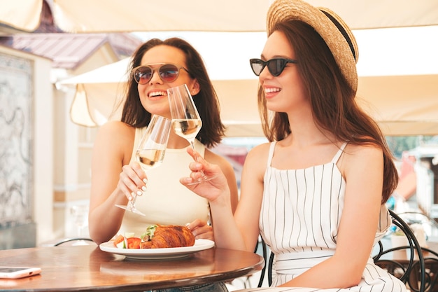 Две молодые красивые улыбающиеся женщины-хипстеры в модной летней одеждеБеззаботные женщины позируют в кафе на веранде на улицеПозитивные модели пьют белое виноЕдят хАкруассан с овощамиГоворя