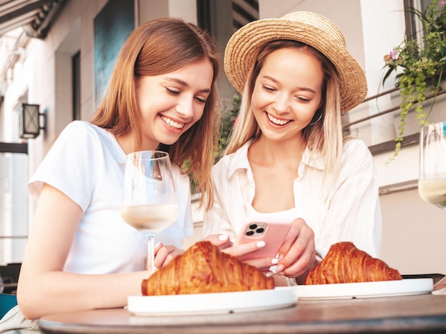 Two young beautiful smiling hipster female in trendy summer clothesCarefree women sitting at veranda cafe in the streetPositive models drinking white wineEating croissantLooking at phone screen