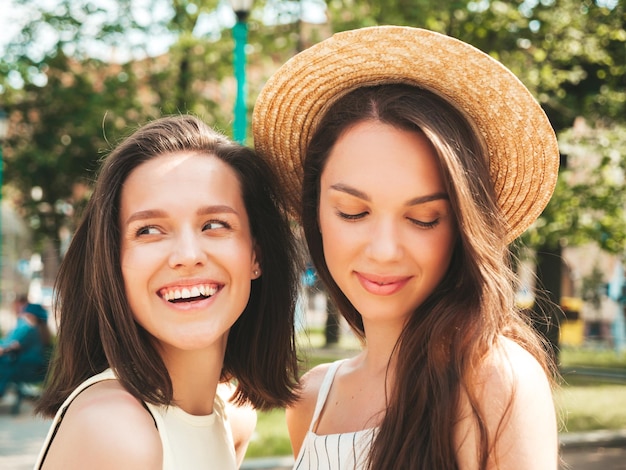 Two young beautiful smiling hipster female in trendy summer clothesSexy carefree women posing on the street background in hat Positive pure models having fun at sunset hugging