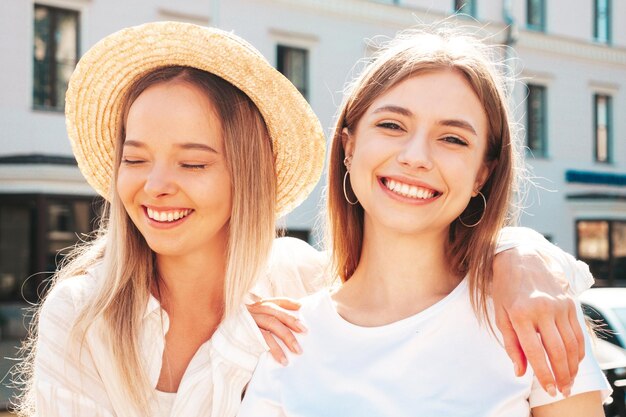 トレンディな夏の服を着た2人の若い美しい笑顔のヒップスターの女性帽子をかぶって通りでポーズをとるセクシーな屈託のない女性日没で楽しんでいるポジティブな純粋なモデル