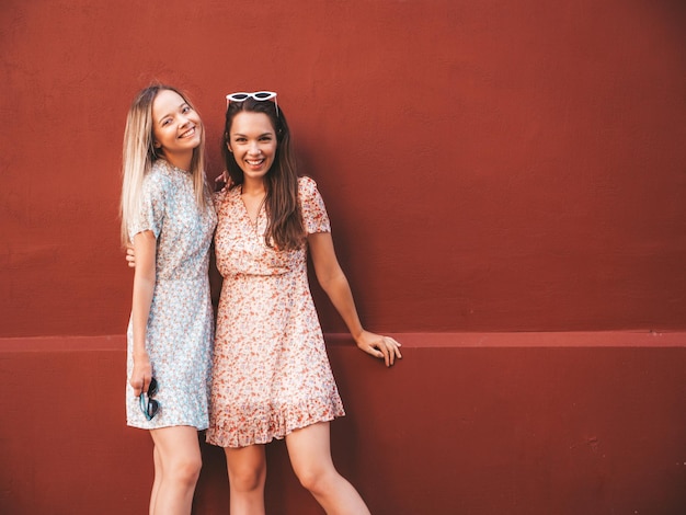 Две молодые красивые улыбающиеся хипстерши в модных летних платьяхСексуальные беззаботные женщины позируют на фоне улицы возле красной стены Позитивные чистые модели веселятся на закате, обнимаются