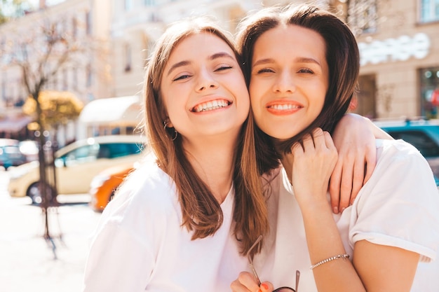 Two young beautiful smiling hipster female in trendy summer white t-shirt clothes and jeans.Sexy carefree women posing on the street background. Positive models having fun, hugging and going crazy