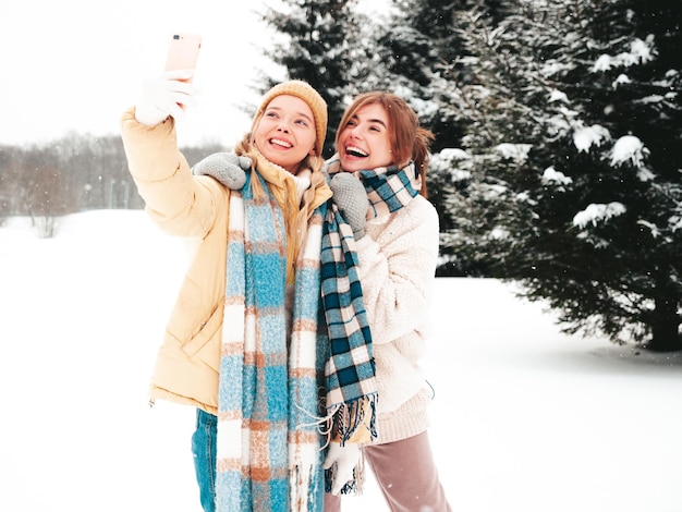 Two young beautiful smiling hipster female in trendy warm clothes and scarfs.Carefree women posing in the street in park. Positive pure models having fun in snow. Enjoying winter moments.Taking selfie