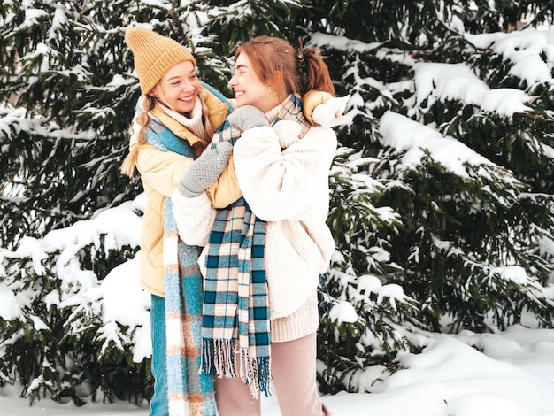 Two young beautiful smiling hipster female in trendy warm clothes and scarfs.Carefree women posing in the street in park. Positive pure models having fun in snow. Enjoying winter moments. Xmas concept